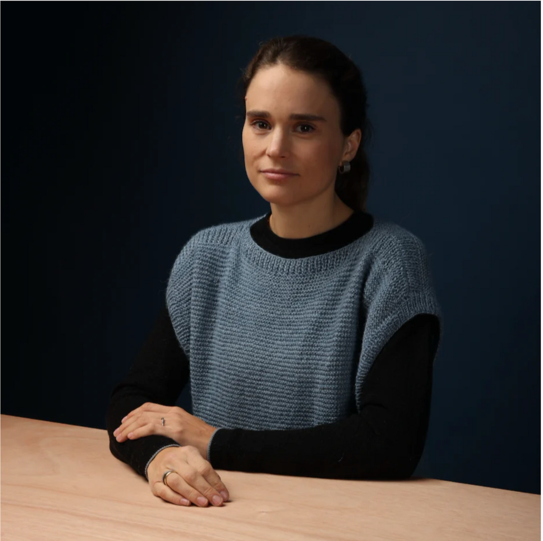 A professional photo of Florencia Hurtado, Creative Director of Selvedge Magazine. She is sat at a wooden table, against a dark background.